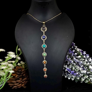 May include: A long gold chain necklace with seven round gemstone beads in different colors. The beads are set in gold-toned bezels and are arranged in a vertical line. The necklace is displayed on a black mannequin.