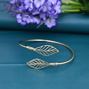 May include: A gold-colored, open cuff bracelet with leaf-shaped accents. The bracelet features two openwork leaf designs, one at each end, connected by a slender, curved band. The bracelet is displayed on a teal surface, with purple flowers in the background.
