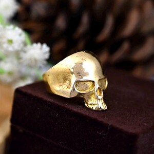 May include: A gold skull ring with a detailed design. The ring is on a brown velvet surface.