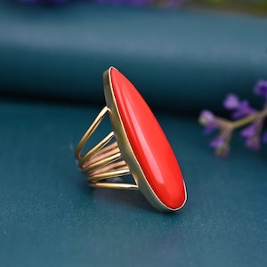 May include: A gold-toned ring with a vibrant red, elongated oval stone. The ring features a multi-band design on the band, adding a unique touch. The red stone is the focal point, set in a gold frame, creating a striking contrast.