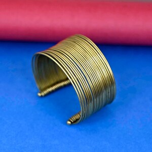 May include: A gold-toned metal bangle bracelet made of multiple thin, stacked bands. The bracelet is open and has a curved shape.