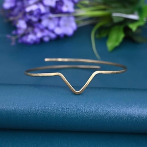 May include: A gold-colored, open cuff bracelet with a V-shaped design. The bracelet is made of a thin, hammered metal and sits on a blue surface. The background includes purple flowers and green leaves.
