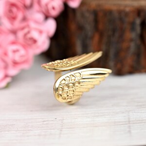 K&ouml;nnte beinhalten: Ein goldfarbener Ring mit einem Fl&uuml;geldesign. Der Ring hat zwei detaillierte Fl&uuml;gel, einen auf jeder Seite, mit strukturierten Federdetails. Der Ring befindet sich vor einem verschwommenen Hintergrund mit rosa Blumen und einer Holzoberfl&auml;che.