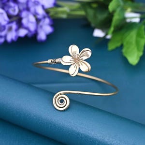May include: Gold-toned arm cuff featuring a floral design. The cuff has a spiral detail at one end and a flower at the other. The jewelry is set against a teal background with purple flowers and green leaves.