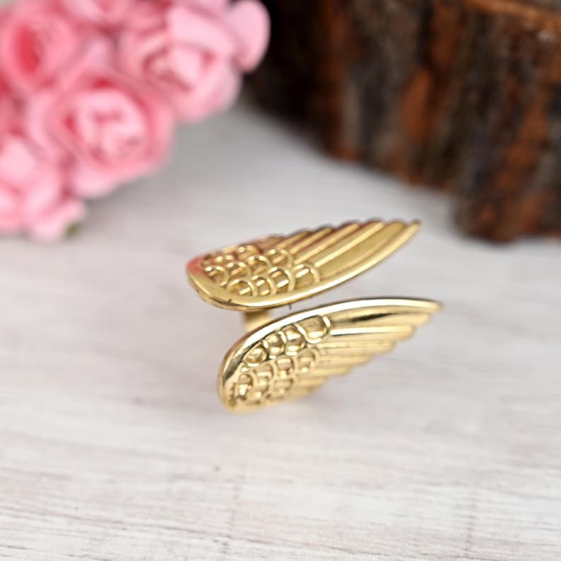 K&ouml;nnte beinhalten: Ein goldfarbener Ring mit Fl&uuml;geldesign. Der Ring hat zwei detaillierte Fl&uuml;gel, jeder mit strukturierten Mustern, die einen geschichteten Effekt erzeugen. Der Ring befindet sich auf einer hellen Holzoberfl&auml;che mit rosa Blumen im Hintergrund.