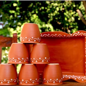 May include: A terracotta clay tea set featuring six small, stacked cups and a matching tray. The cups and tray are a warm orange-brown color and decorated with a white dotted wave pattern around the rims.