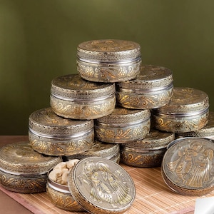 Puede incluir: Una pila de recipientes redondos de metal con acabados en oro y plata, con intrincados grabados florales. Un recipiente abierto en primer plano revela anacardos, mientras que su tapa muestra un relieve de dos figuras.