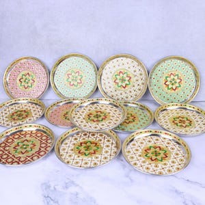 May include: A collection of eleven round metal plates featuring gold rims and intricate, colourful floral patterns in the centre. Each plate displays a unique combination of pastel and bold colours, including pink, green, white, and red, with geometric motifs.