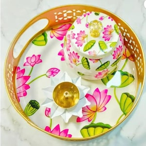 以下が含まれることがあります： 金色の縁とハンドルが付いた装飾トレイ。ピンクの蓮の花と緑の葉の白い花柄。お揃いの蓋付きジャーと、金色の中心部を備えた白い蓮の形をしたキャンドルホルダーがトレイにあります。