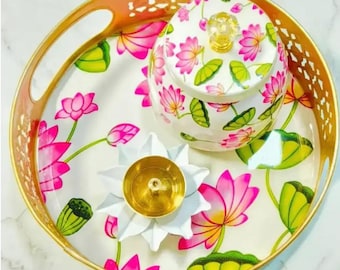 May include: A decorative tray with a gold rim and handles, featuring a white floral design with pink lotus flowers and green leaves. A matching lidded jar and a white lotus-shaped candle holder with a gold center are on the tray.