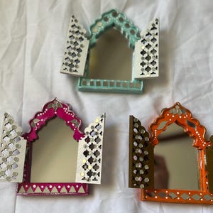 Könnte beinhalten: Drei dekorative Spiegel mit verzierten Rahmen. Die Spiegel sind in Türkis, Rosa und Orange lackiert. Jeder Spiegel hat ein weißes Gitterdesign an den Seiten.