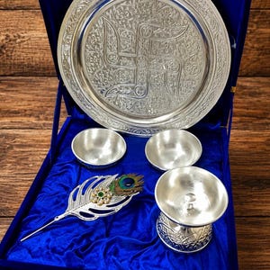Puede incluir: Un conjunto de metal plateado que incluye un plato circular con una esvástica grabada, dos cuencos pequeños, una copa con pedestal y un accesorio en forma de pluma con una piedra verde, presentado en una caja forrada de terciopelo azul.