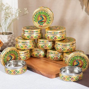 May include: A collection of round metal containers with gold-toned exteriors, featuring hand-painted peacock motifs in green, blue, and red. Each container has a removable lid and a polished silver-toned interior, stacked in a pyramid arrangement.