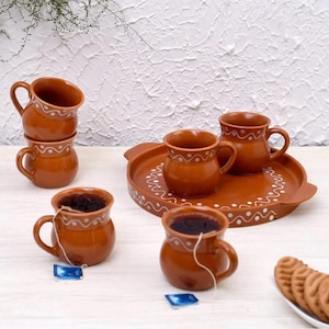 May include: A terracotta-coloured ceramic tea set, including mugs and a serving tray. The mugs feature a rounded shape, handles, and a white dotted pattern. Tea bags are visible in two mugs. A plate of biscuits is in the background.