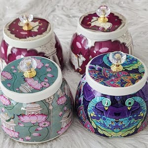 May include: Four decorative ceramic jars with lids. The jars have a round shape and are adorned with intricate patterns and designs. Each jar features a crystal knob on the lid. The jars are in shades of teal, purple, and burgundy.