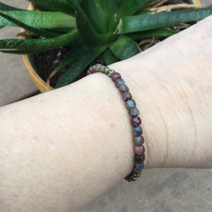 May include: A bracelet made with small, faceted gemstone beads in shades of red, gray, and blue. The beads are strung on a thin, gold-colored wire.