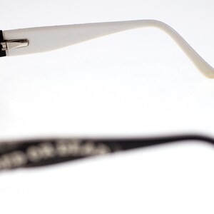May include: Black-framed spectacles with white arms. The glasses have a black frame around the lenses and white arms that curve to the ears. The background is white.