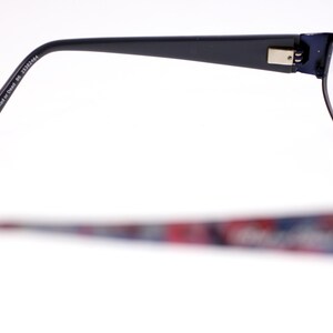 May include: Eyeglasses with a dark blue frame. The upper frame is in focus, showing the arm with the text "52019-135 Red or Dead BC 25383664". The lower frame is out of focus, with a colourful pattern.