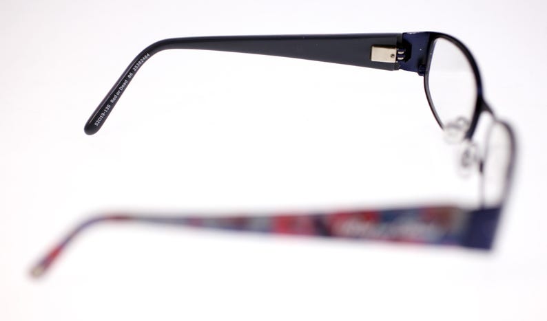 May include: Eyeglasses with a dark blue frame. The upper frame is in focus, showing the arm with the text "52019-135 Red or Dead BC 25383664". The lower frame is out of focus, with a colourful pattern.
