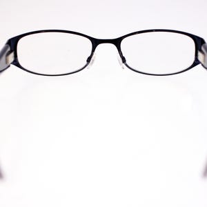 May include: Black-framed spectacles with oval lenses. The glasses have a thin, dark frame and clear lenses. The temples are dark and extend outwards. The spectacles are set against a white background.
