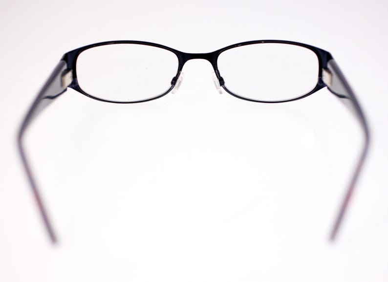 May include: Black-framed spectacles with oval lenses. The glasses have a thin, dark frame and clear lenses. The temples are dark and extend outwards. The spectacles are set against a white background.