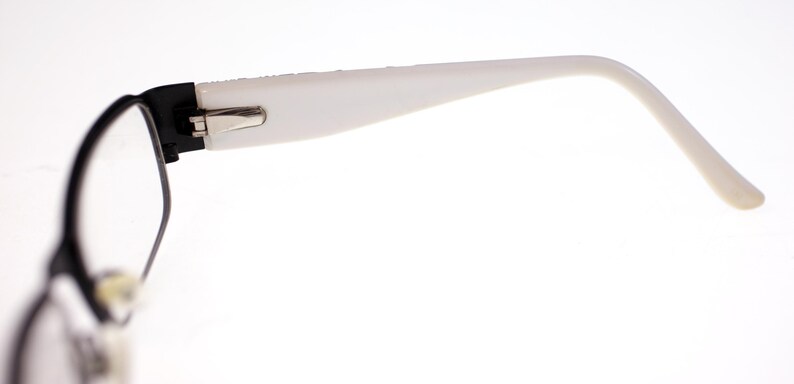 May include: Close-up of eyeglasses with a black frame and white temples. The glasses have a rectangular lens shape and a silver hinge. The white temples curve gently, ending in a rounded tip. The background is plain white.
