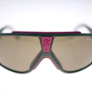 May include: Grey sunglasses with brown lenses and a pink bridge detail. The sunglasses have a sporty design and a logo on the bridge. The lenses are light brown, and the frame is grey.