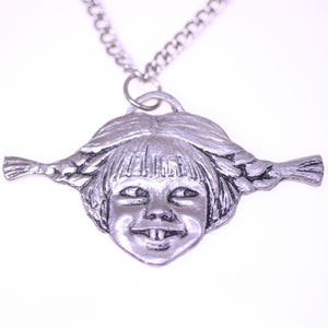 May include: Silver-toned pendant necklace featuring a smiling face with pigtails. The pendant is a detailed, three-dimensional design of a child's face with a wide grin and braided hair, suspended from a silver chain.