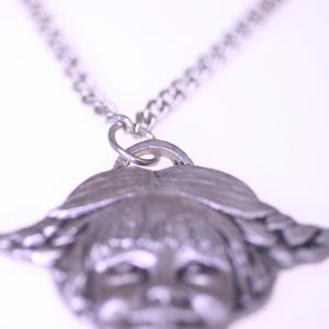 May include: A silver-toned pendant necklace featuring a detailed face with braided hair. The pendant is attached to a silver chain. The necklace is a vintage style accessory.