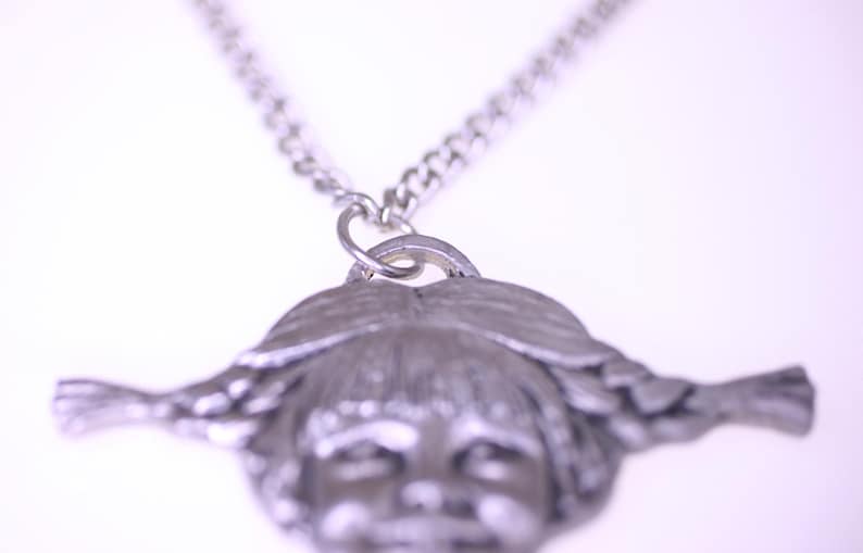 May include: A silver-toned pendant necklace featuring a detailed face with braided hair. The pendant is attached to a silver chain. The necklace is a vintage style accessory.