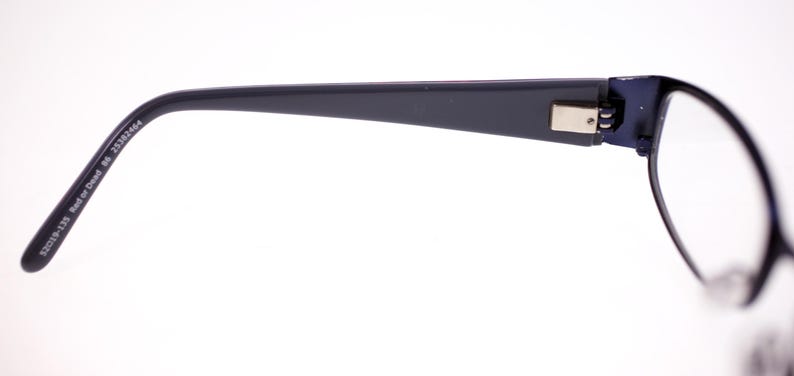 May include: Dark blue eyeglasses with a rectangular frame and a silver hinge. The temple arm is marked with text. The lenses are clear. The glasses are set against a white background.