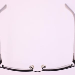 May include: Black-framed spectacles with a rectangular shape, featuring a thin, sleek design. The temples are straight and extend to the ear pieces. The nose pads are white. The glasses are set against a soft, light background.