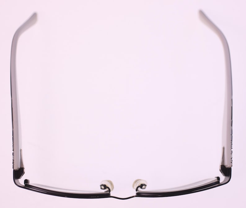 May include: Black-framed spectacles with a rectangular shape, featuring a thin, sleek design. The temples are straight and extend to the ear pieces. The nose pads are white. The glasses are set against a soft, light background.