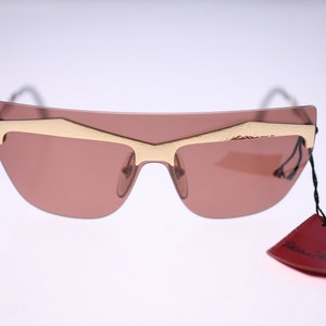 Può includere: Occhiali da sole dorati con lenti marroni e un'etichetta rossa con la scritta "Prada".