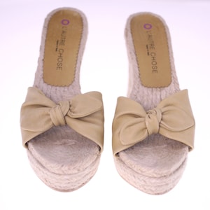 Puede incluir: Un par de sandalias de cuña beige con un gran lazo en la parte delantera. Las sandalias están hechas de un material tejido y tienen una parte superior de cuero. El nombre de la marca "L'AUTRE CHOSE" está impreso en la plantilla.