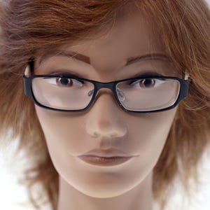 May include: Close-up of black rectangular eyeglasses with clear lenses. The frames have a modern, thick design. The glasses are set against a neutral background, highlighting their sleek appearance. The image focuses on the eyewear.