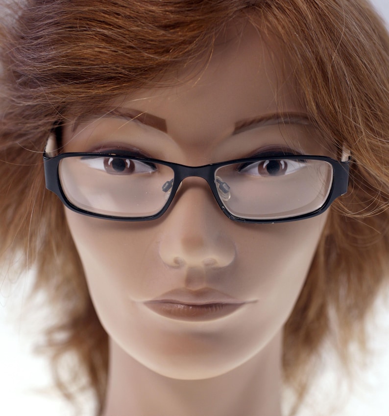 May include: Close-up of black rectangular eyeglasses with clear lenses. The frames have a modern, thick design. The glasses are set against a neutral background, highlighting their sleek appearance. The image focuses on the eyewear.