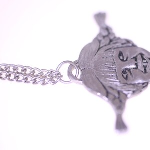 May include: A silver-toned pendant necklace featuring a detailed face design. The pendant is attached to a delicate chain. The face has intricate details, and the overall design is vintage-inspired.