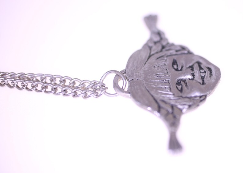 May include: A silver-toned pendant necklace featuring a detailed face design. The pendant is attached to a delicate chain. The face has intricate details, and the overall design is vintage-inspired.