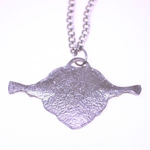 May include: A silver-colored pendant necklace. The pendant has a textured, irregular shape with two extended wing-like features. The chain is a delicate, linked design. The necklace is set against a white background.