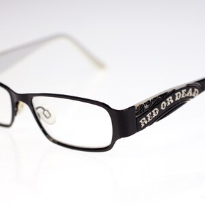 May include: Black rectangular eyeglasses with clear lenses. The frames are black with white arms. The words "RED OR DEAD" are printed in white on the arms. The glasses are set against a white background.