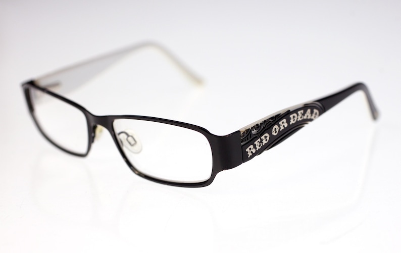 May include: Black rectangular eyeglasses with clear lenses. The frames are black with white arms. The words "RED OR DEAD" are printed in white on the arms. The glasses are set against a white background.