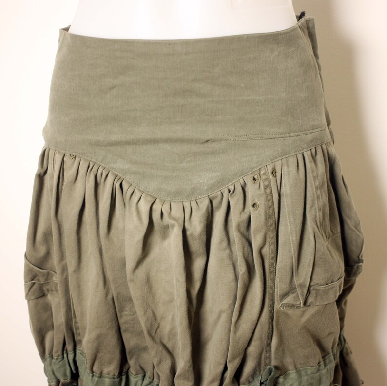 May include: A green utility skirt with multiple pockets and a gathered, tiered design. The skirt is made of a durable, canvas-like fabric.