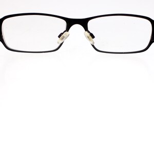 May include: Black rectangular spectacles with clear lenses. The frames are black with white arms. The glasses are set against a bright white background.