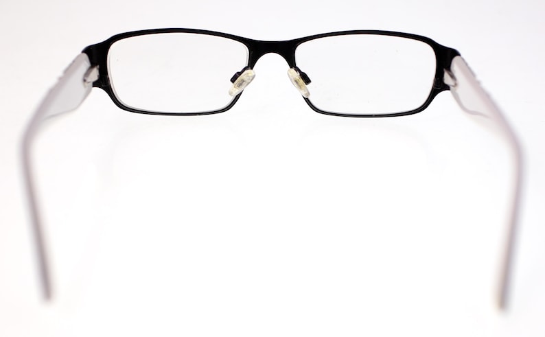 May include: Black rectangular spectacles with clear lenses. The frames are black with white arms. The glasses are set against a bright white background.