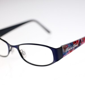 May include: Navy blue eyeglasses with a rectangular frame. The temples have a colourful floral pattern and the text "Red or Dead". The lenses are clear.