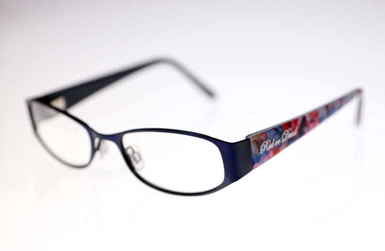 May include: Navy blue eyeglasses with a rectangular frame. The temples have a colourful floral pattern and the text "Red or Dead". The lenses are clear.