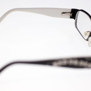 May include: Black and white spectacles with a rectangular frame and clear lenses. The arms are white, and the frame is black with decorative detailing. The glasses are designed for vision correction.