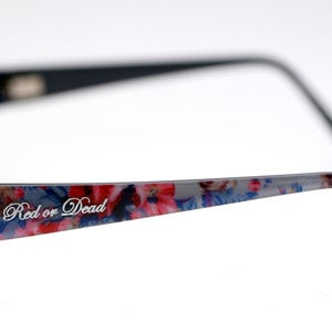 May include: Black framed eyeglasses with a floral pattern on the temples. The text "Red or Dead" is printed on the temple. The frames are black with a rectangular shape.