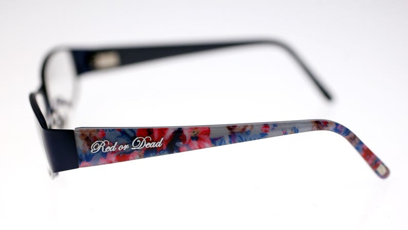May include: Black framed eyeglasses with a floral pattern on the temples. The text "Red or Dead" is printed on the temple. The frames are black with a rectangular shape.